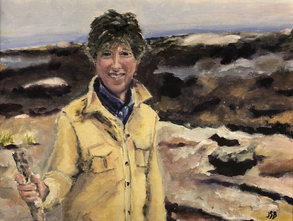 Oil painting portrait of a woman with short brown hear standing on some large rocks at the water's edge. We see only from the waist up. Woman is holding a walking stick. She is wearing a yellowish/tan shirt and is looking at the viewer and smiling.